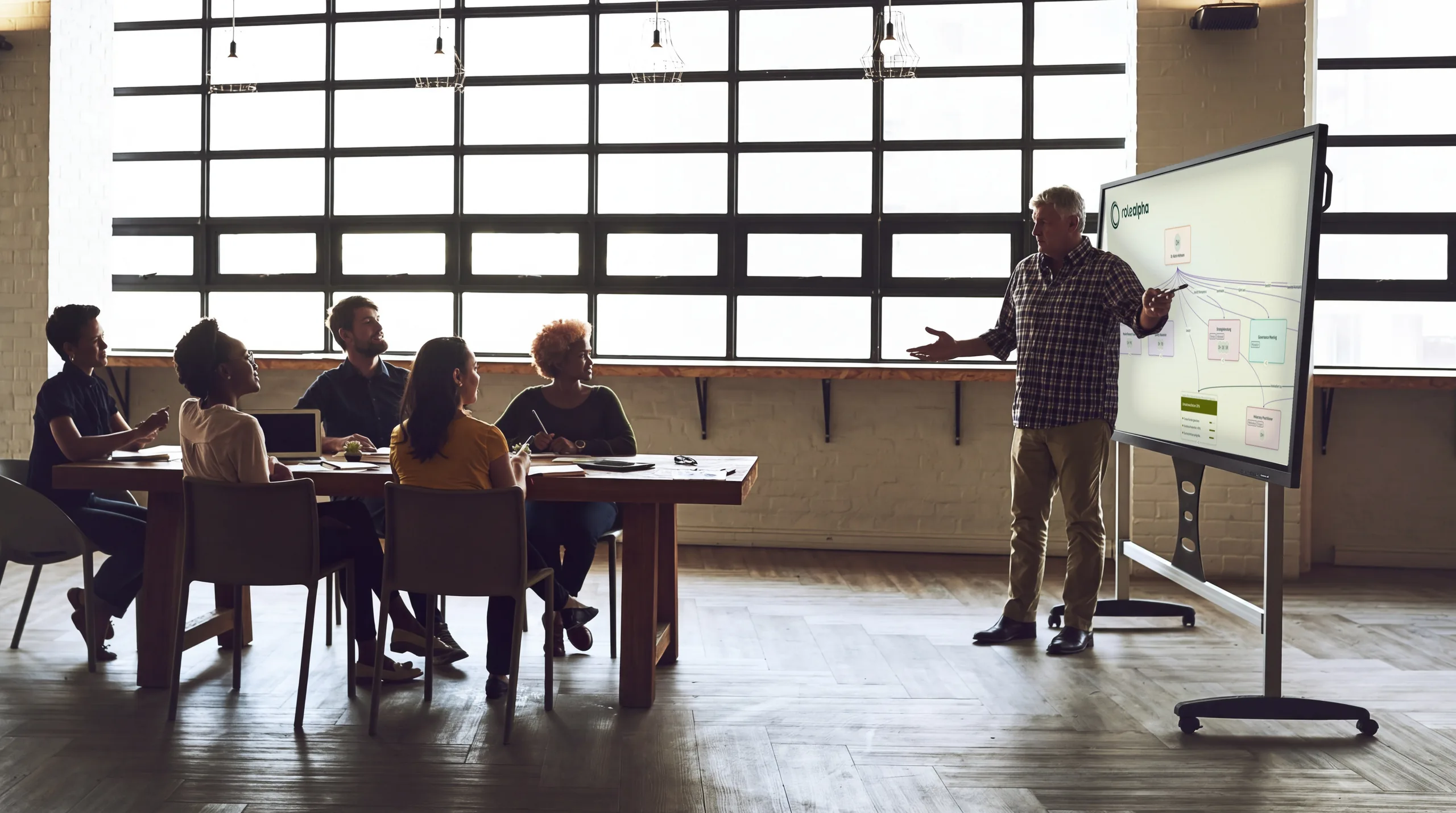 A team sits at a table in a bright loft; a man points at a large display showing the roleALPHA Explorer: connected cards for people, roles, meetings and goals